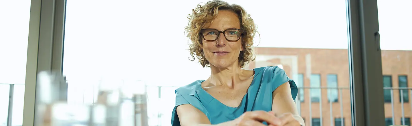 Eine Frau mit Brille sitzt vor einer großen Glasfront an einem Konferenztisch und blickt direkt in die Kamera.