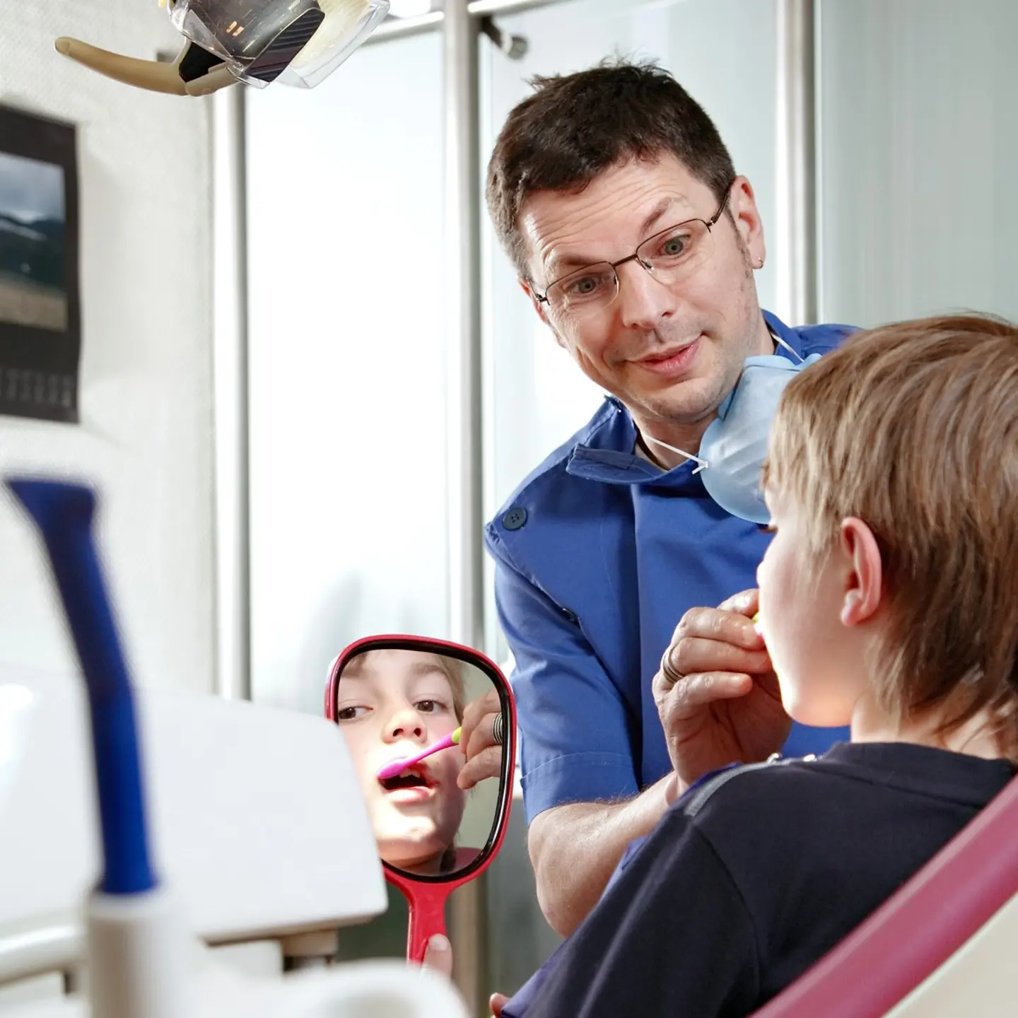 Ein Junge sitzt beim Zahnarzt auf dem Stuhl und schaut in einen Spiegel. Der Arzt zeigt und erklärt ihm etwas.