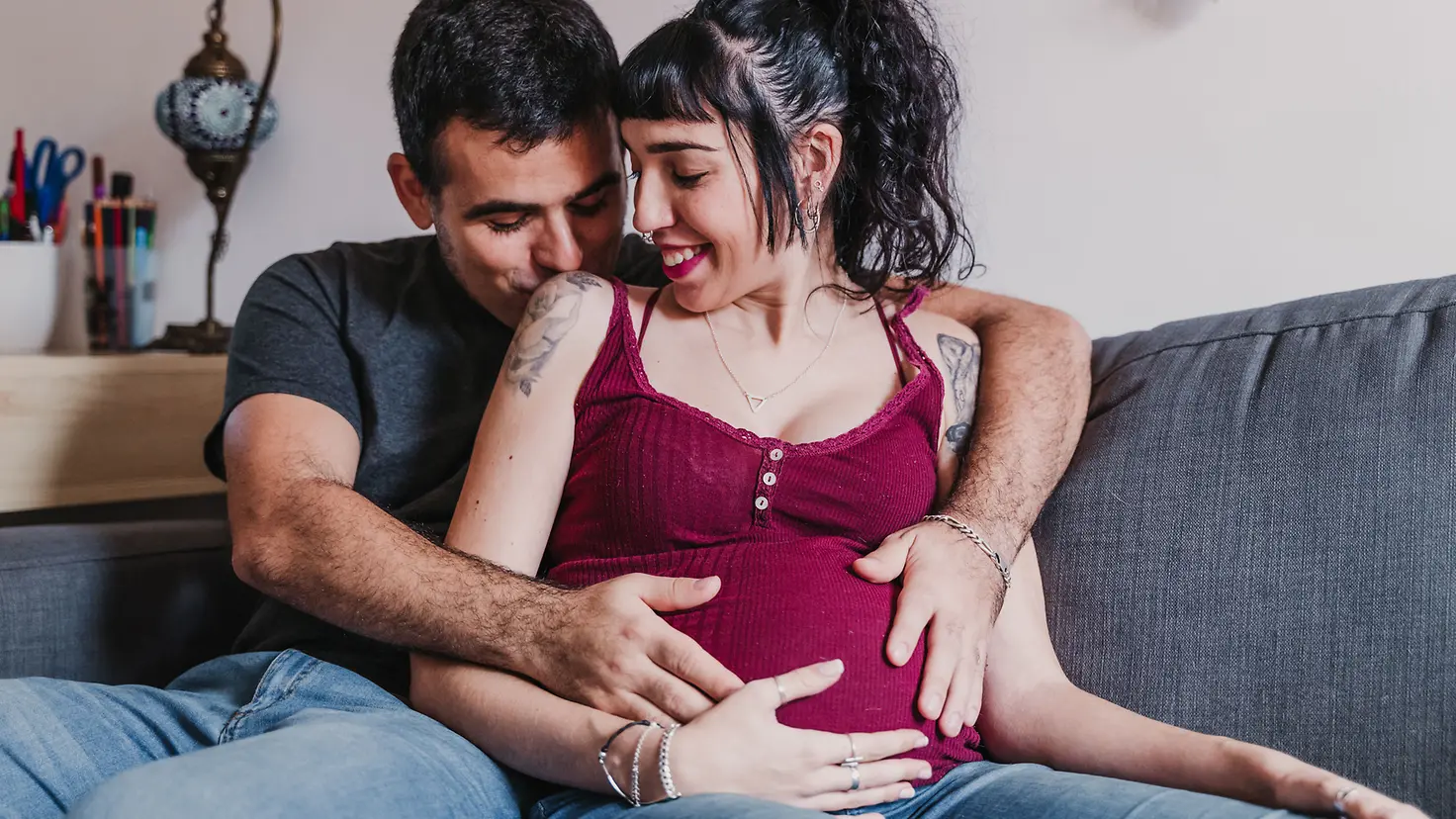 Ein Mann und Frau in der etwa 20. Schwangerschaftswoche sitzen auf einer Couch und halten ihren Babybauch