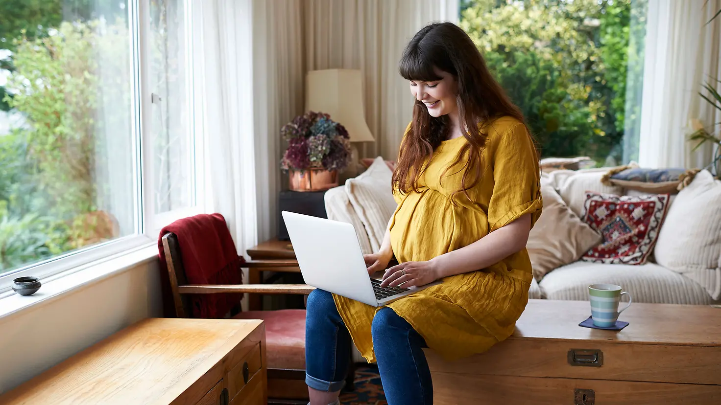 Schwangere Frau um die 33. Schwangerschaftswoche sitzt mit Laptop auf Kommode und lächelt