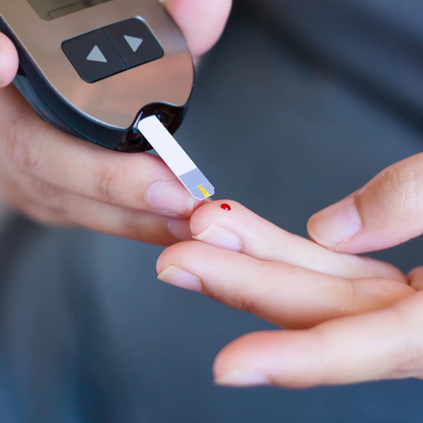 In einem Blutzucker-Messgerät steckt ein Teststreifen. Mit diesem wird ein Bluttropfen von einem Finger der anderen Hand abgenommen.
