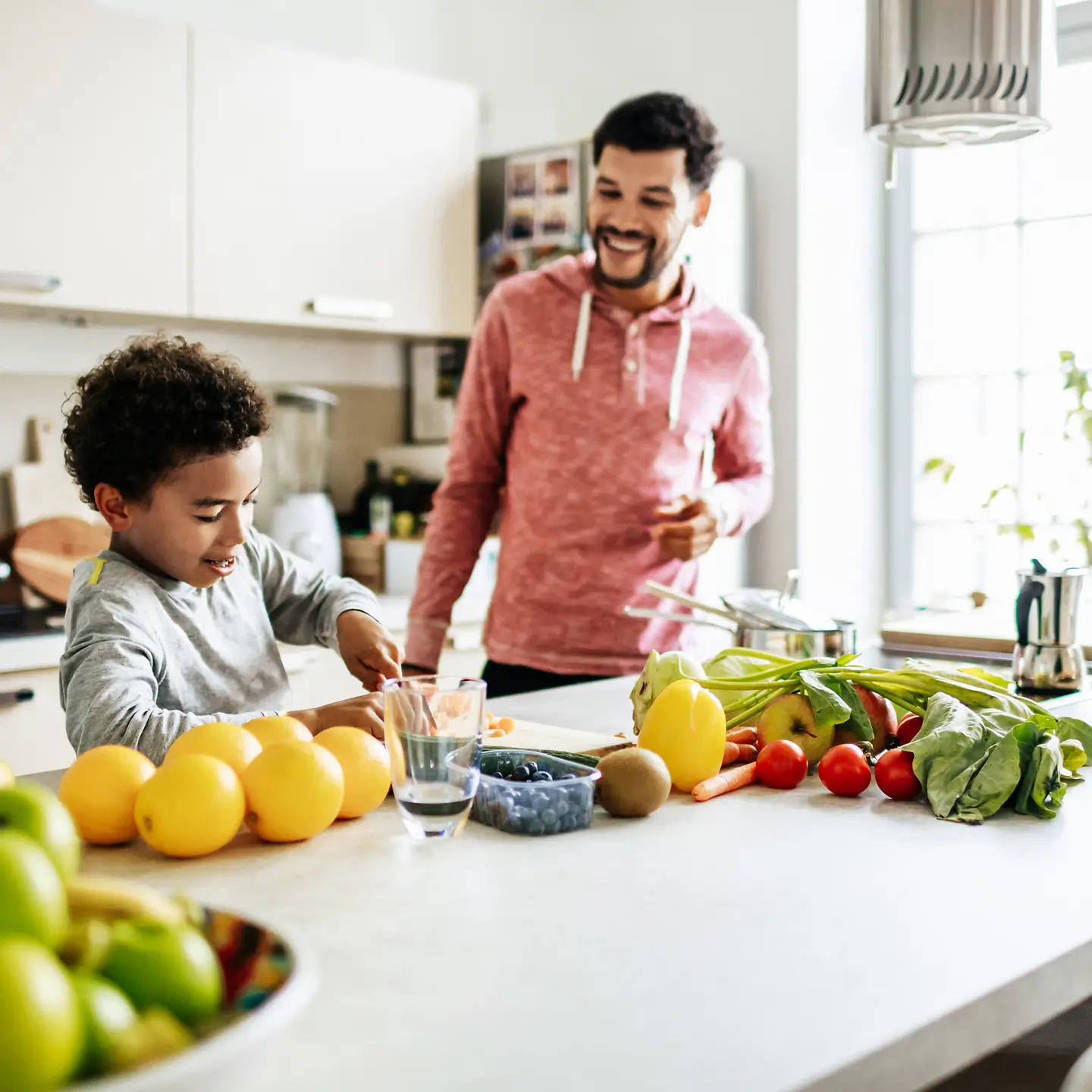 Ein Junge schneidet Obst und Gemüse auf einer Arbeitsplatte in einer Küche. Sein Dad steht daneben und lächelt.