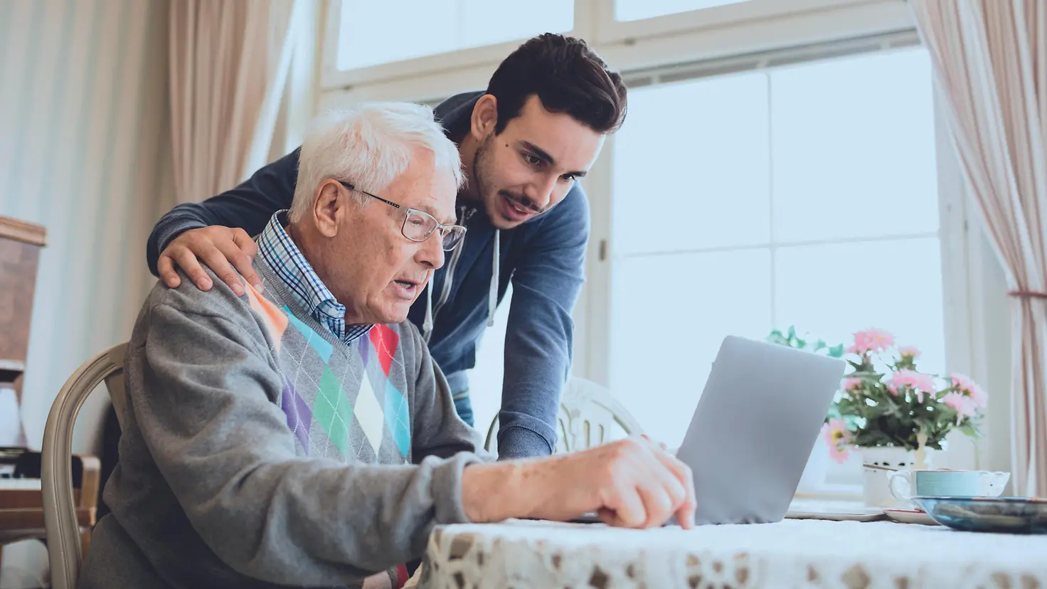 Ein älterer Mann lässt sich von einem Jüngeren etwas an einem Laptop erklären. 
