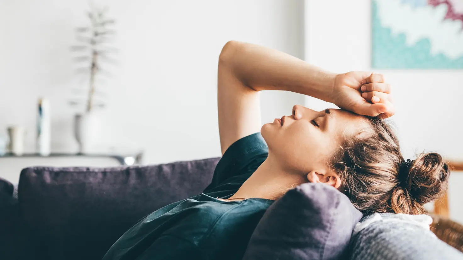 Eine übermüdete Frau liegt mit geschlossenen Augen auf einem Sofa. Sie hält sich mit ihrer rechten Hand die Stirn.