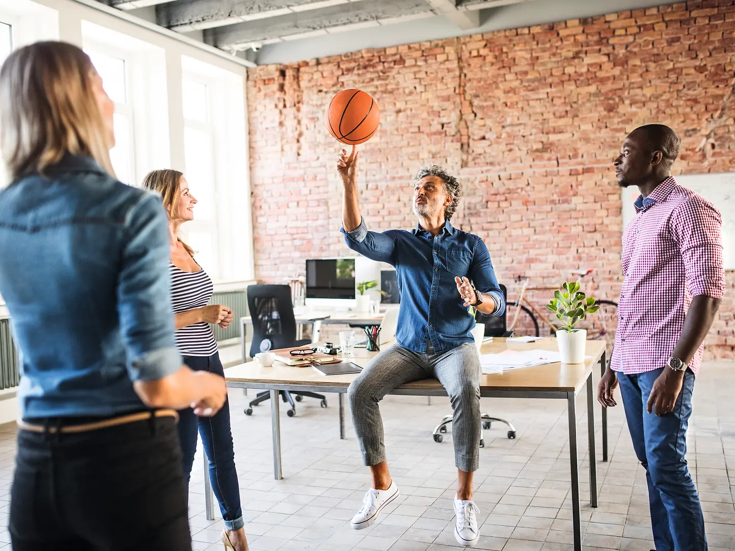 Ein Team steht im Büro. Ein Mitarbeiter sitzt auf dem Tisch und spielt mit einem Basketball.