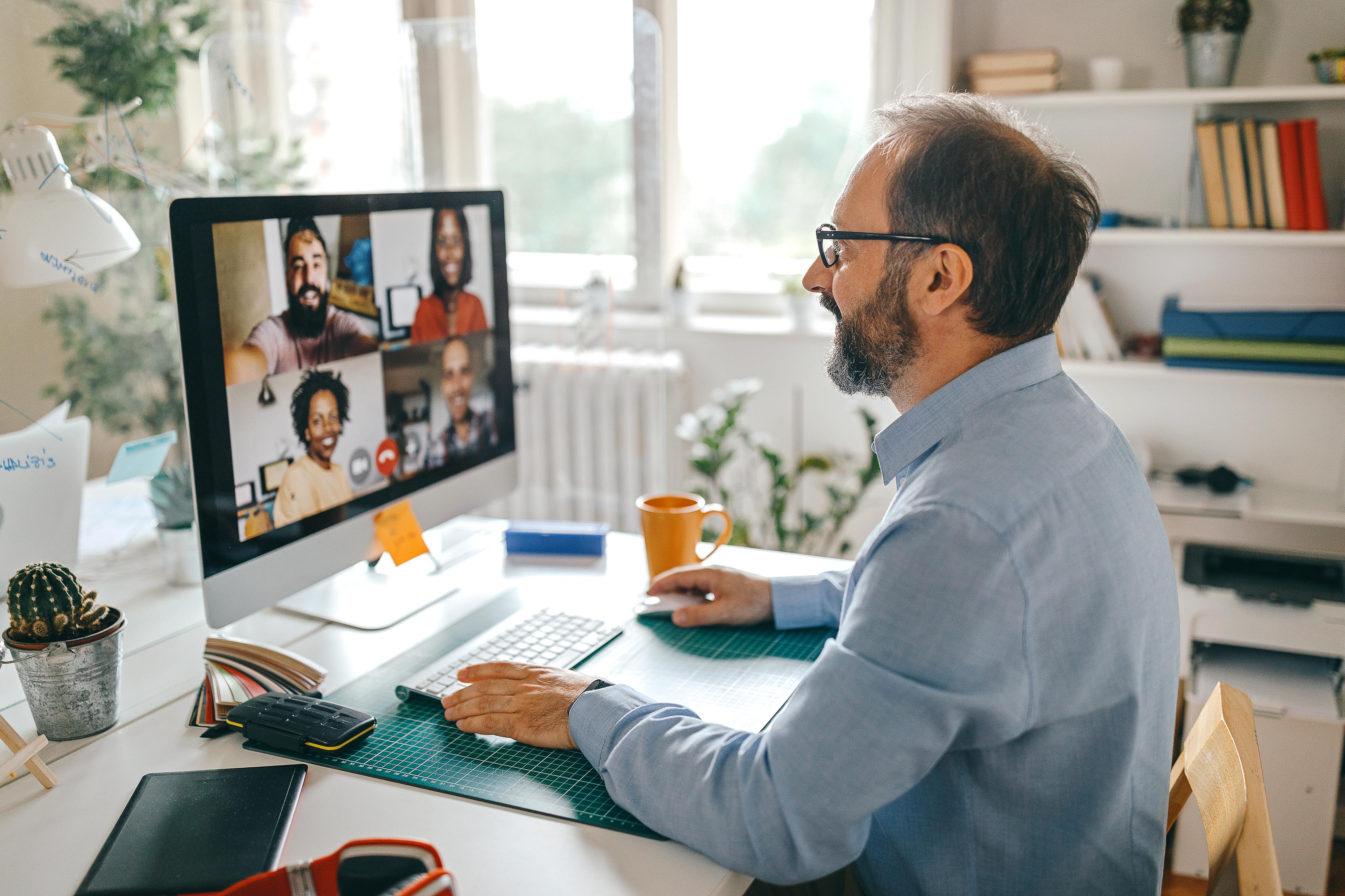 Ein Mann sitzt und schaut auf einen Computerbildschirm, dort ist eine Videokonferenz mit vier lächelnden Teilnehmern zu sehen. 