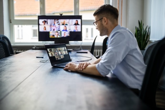 Ein Mann im Hemd sitzt an einem großen Tisch. Vor ihm steht ein Laptop und rechts ein Bildschirm mit einer Videokonferenz.