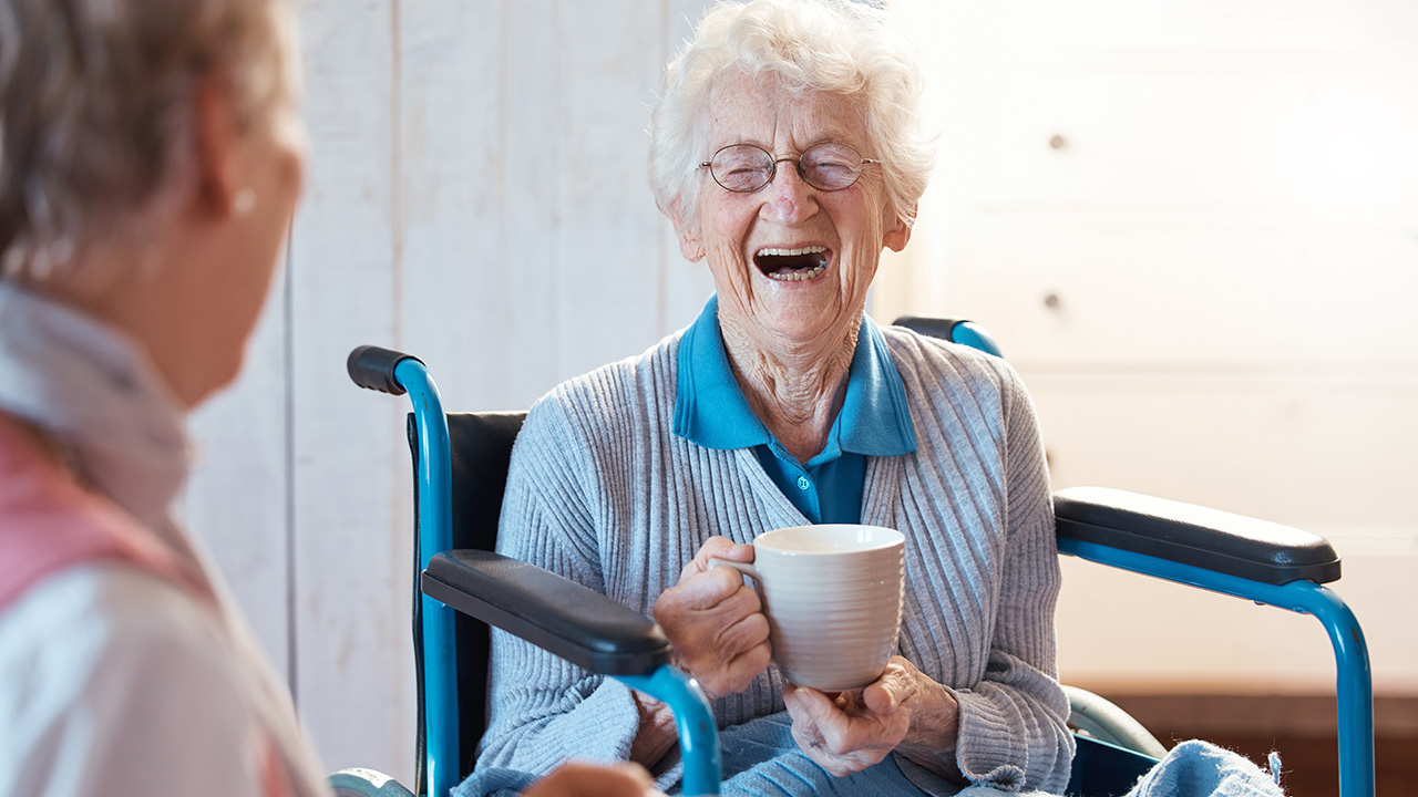 Eine lachende Seniorin im Krankenrollstuhl hält eine Kaffeetasse in der Hand.