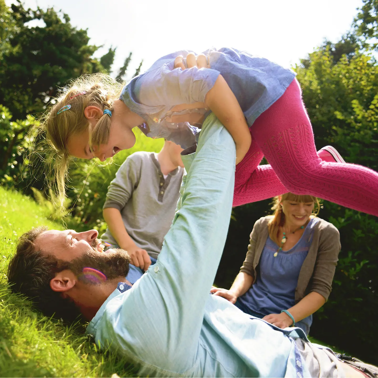 Eine Familie tobt im Garten, der Vater liegt auf dem Rücken und hält seine Tochter hoch