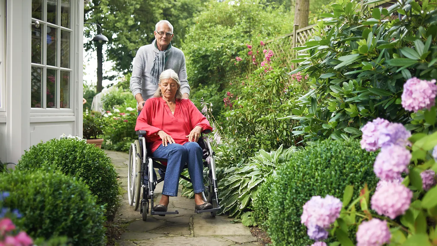 Ein älterer Mann schiebt eine ältere Frau im Rollstuhl in den Garten