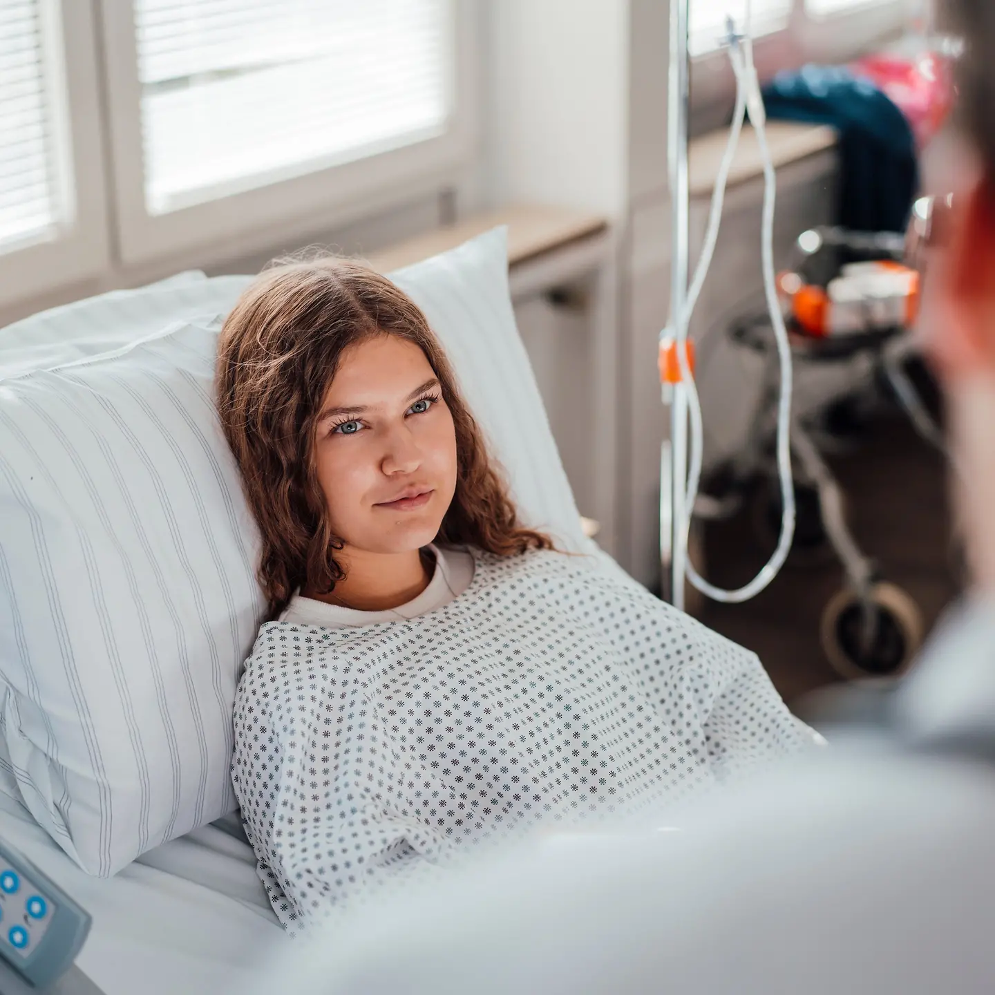 Eine sehr junge Frau liegt in einem Krankehausbett und hört dem Arzt bei ihrer Visite zu.