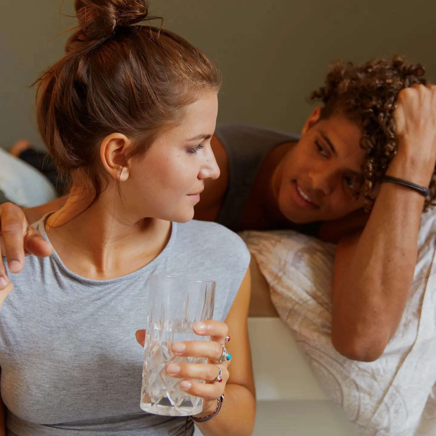 Eine junge Frau trinkt ein Glas Wasser am Bett und ein junger Mann hat den Arm um sie gelegt
