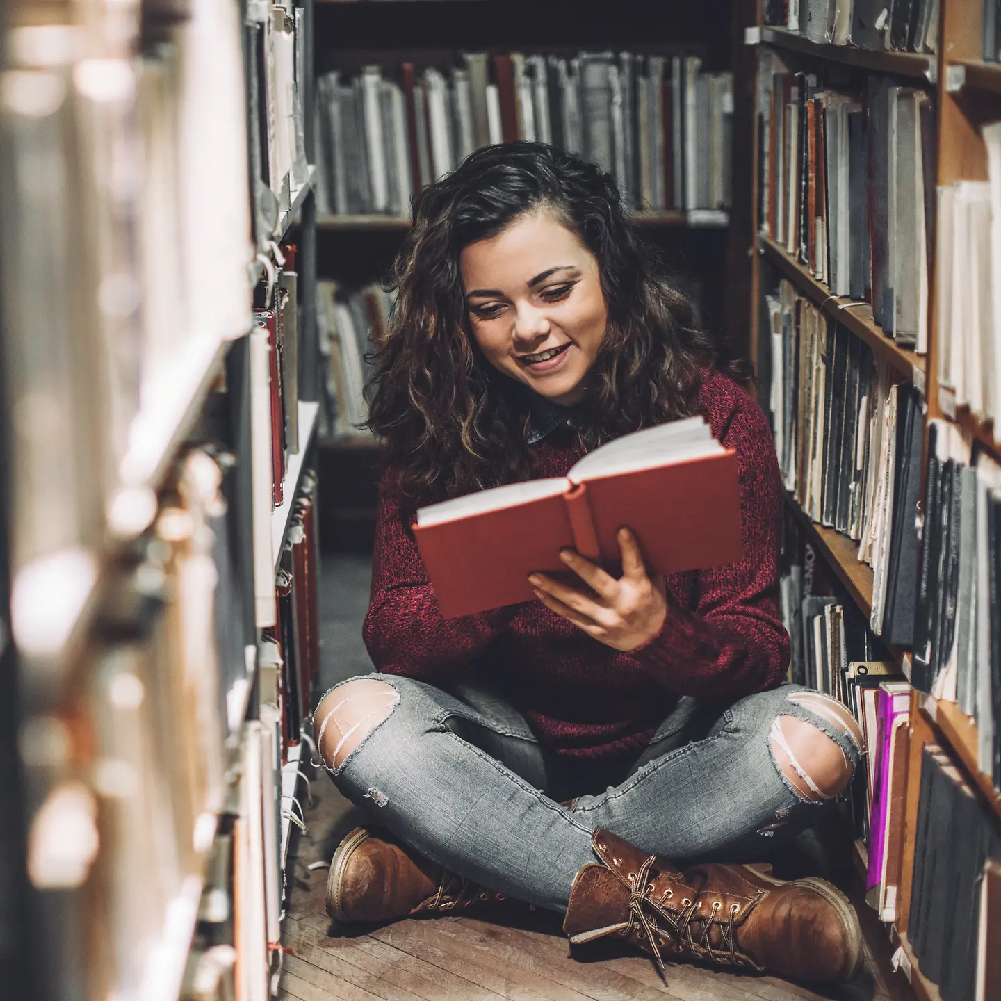 Junge Frau sitzt auf dem Fußboden zwischen Bücherregalen und liest ein Buch