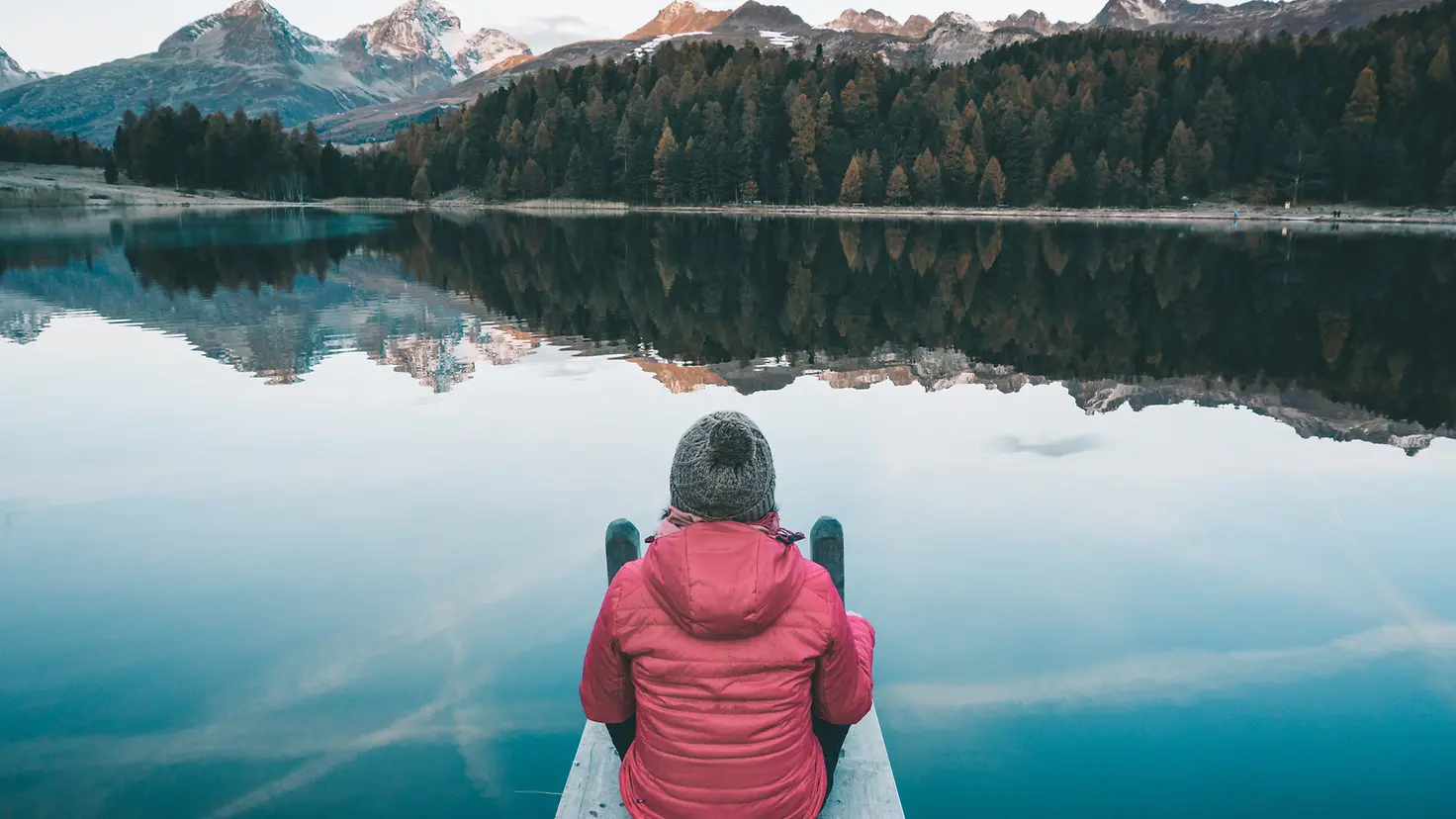 Eine Frau sitzt auf einem Steg am See in den Bergen und schaut in die Ferne
