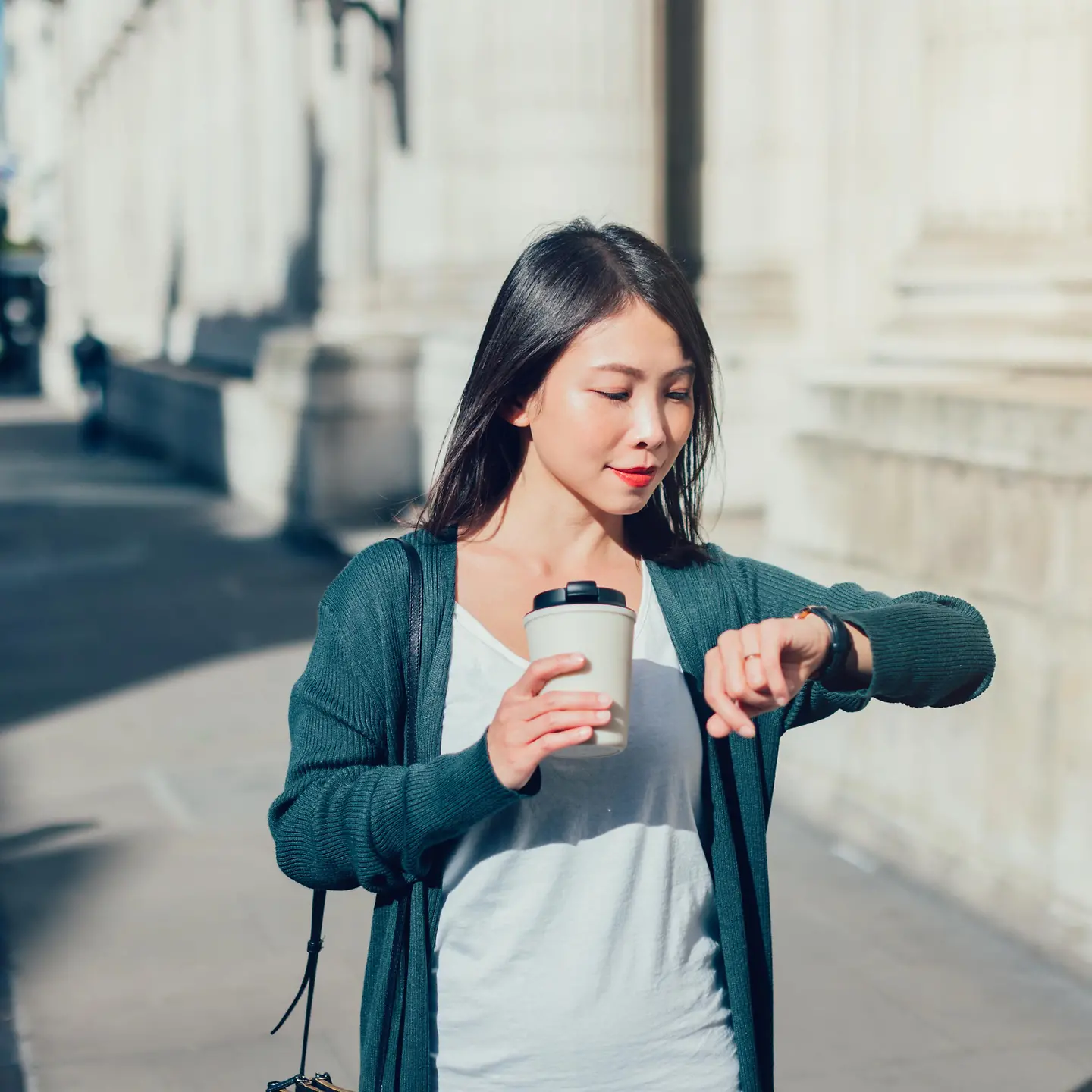 Eine junge Frau mit Kaffeebecher in der Hand schaut auf die Uhr. 