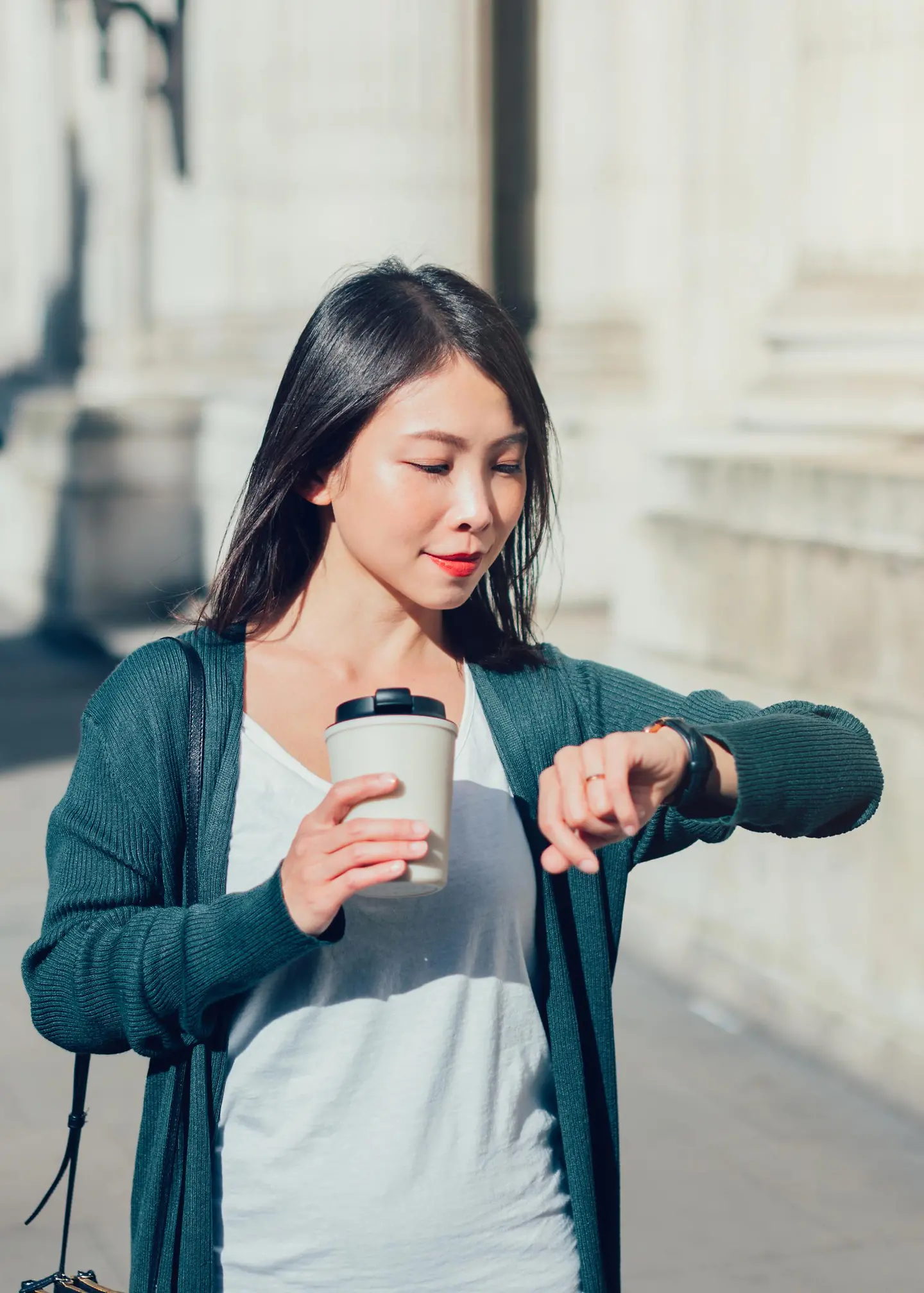 Eine junge Frau mit Kaffeebecher in der Hand schaut auf die Uhr. 