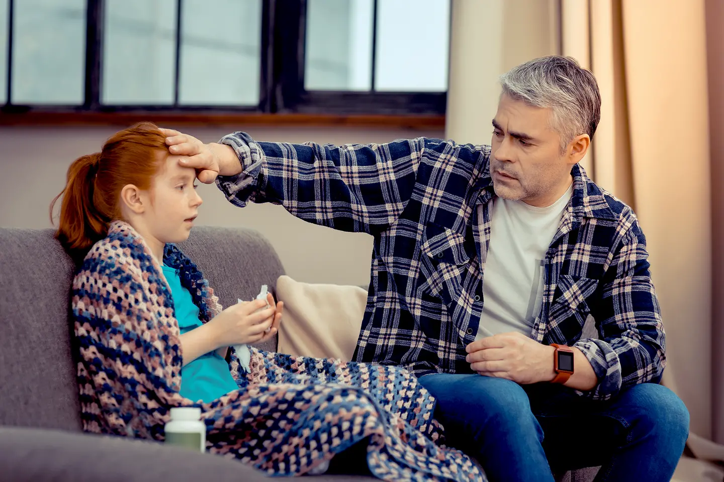 Ein Mädchen liegt krank auf einer Couch und der Vater fühlt ihre Stirn