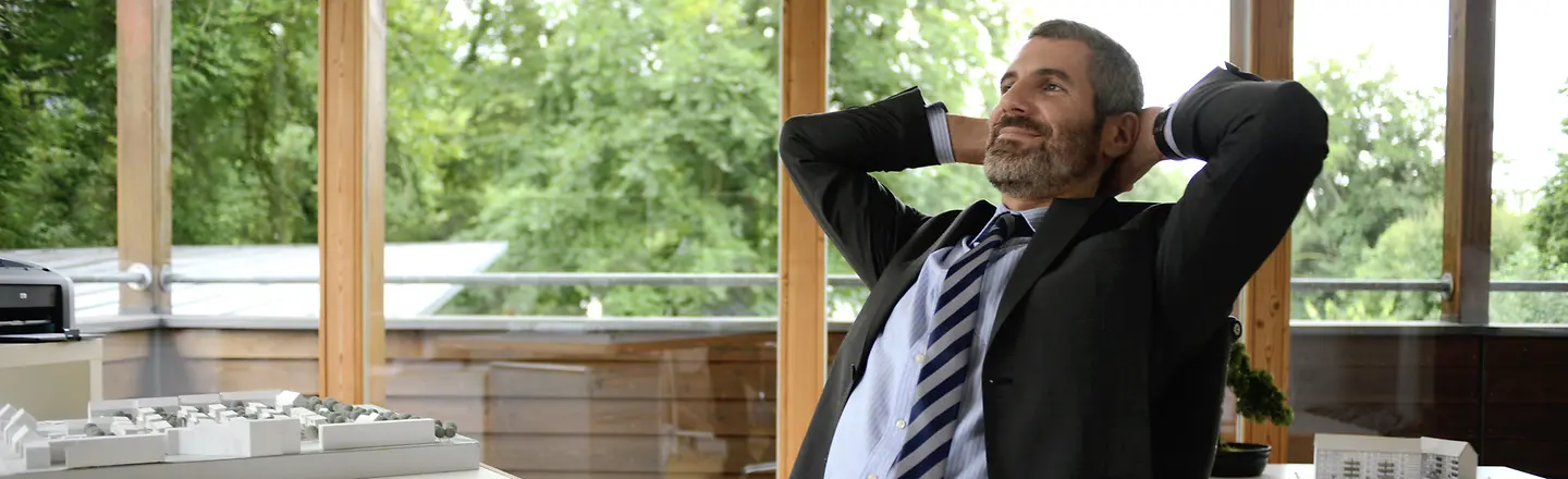 Ein entspannt ausschauender Mann sitzt am Schreibtisch und hat die Hände hinter dem Kopf verschränkt.