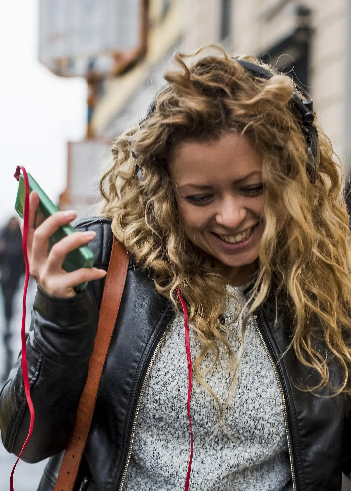 A young woman walks through the street laughing and listening to music on her cell phone.