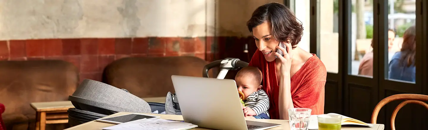 Eine junge Frau sitzt vor einem Laptop im Café, hat ein Baby auf dem Schoß und telefoniert