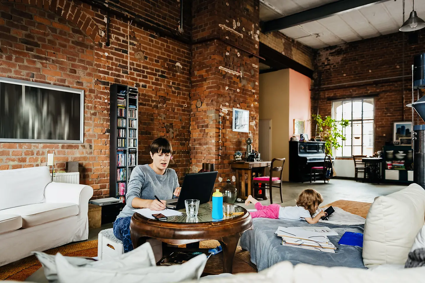 Eine junge Frau arbeitet am Wohnzimmertisch, während ihr Kind auf der Couch spielt