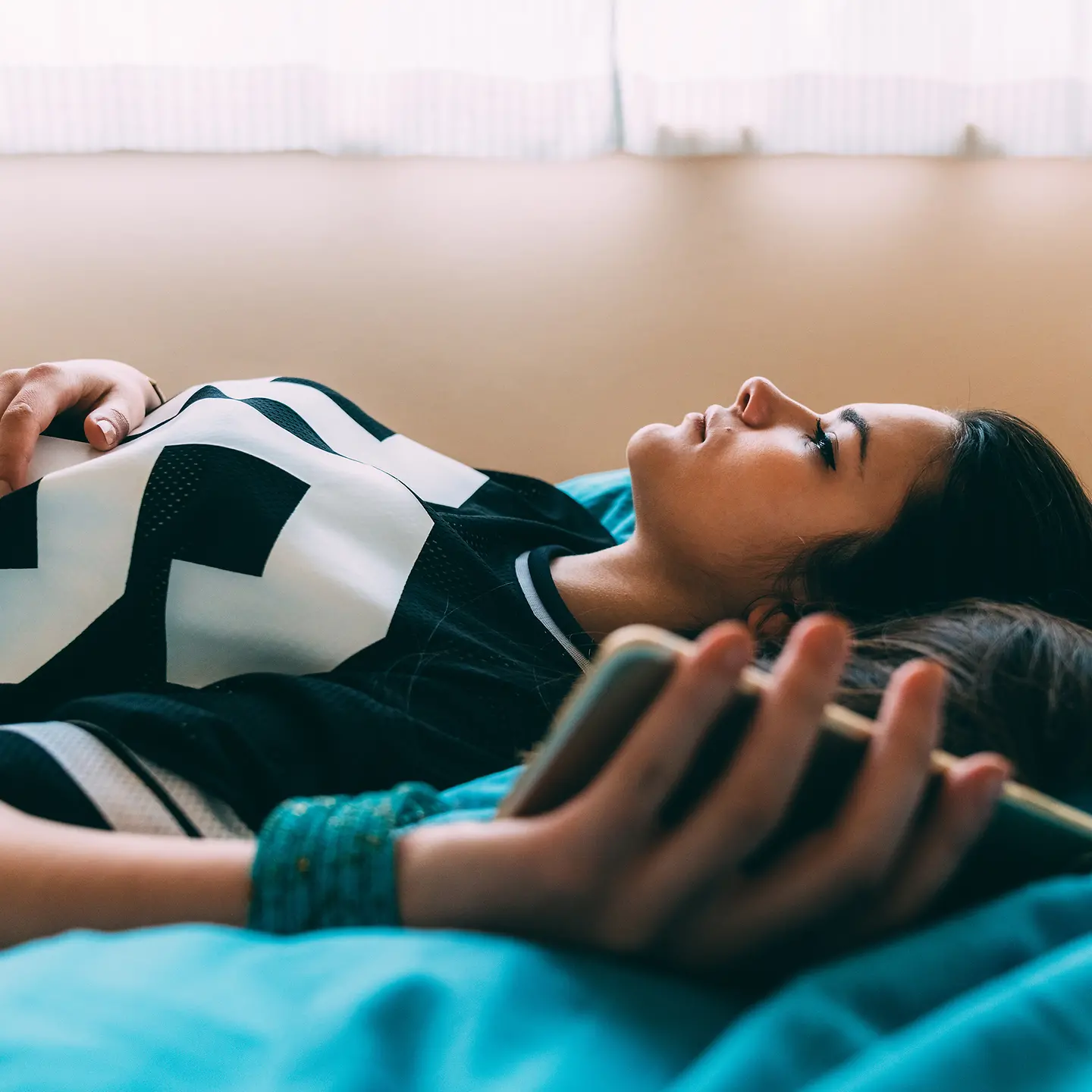 Eine junge Frau liegt auf dem Bett und schaut an die Decke, in der Hand hält sie ein Smartphone