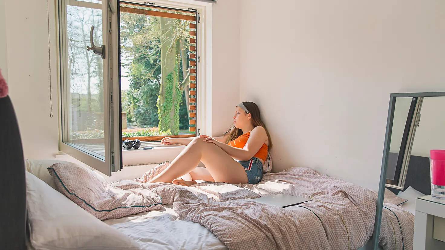 Eine junge Frau sitzt auf dem Bett und schaut nachdenklich aus dem Fenster
