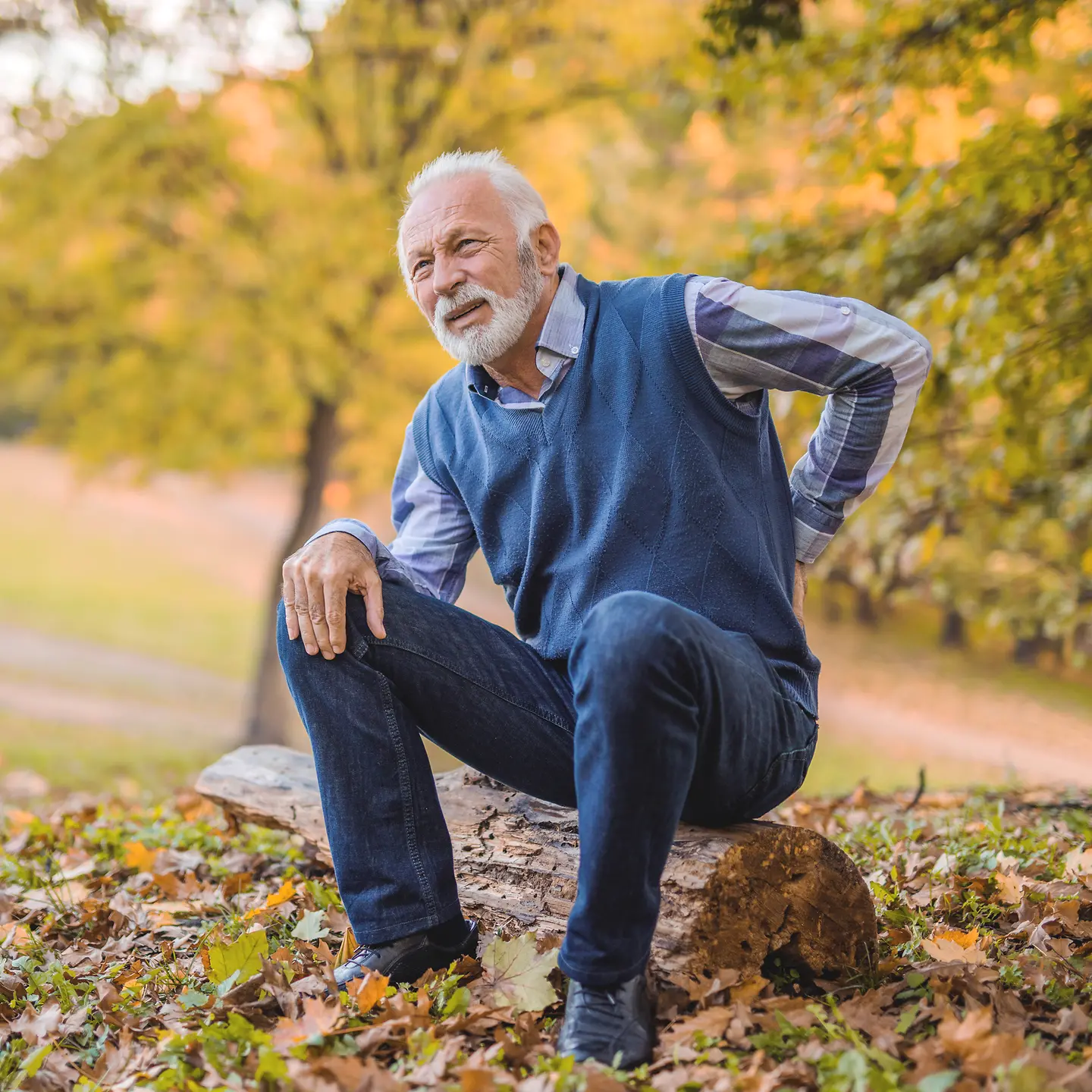 Ein älterer Mann sitzt auf einem Baumstamm und hält sich mit der Hand den Rücken
