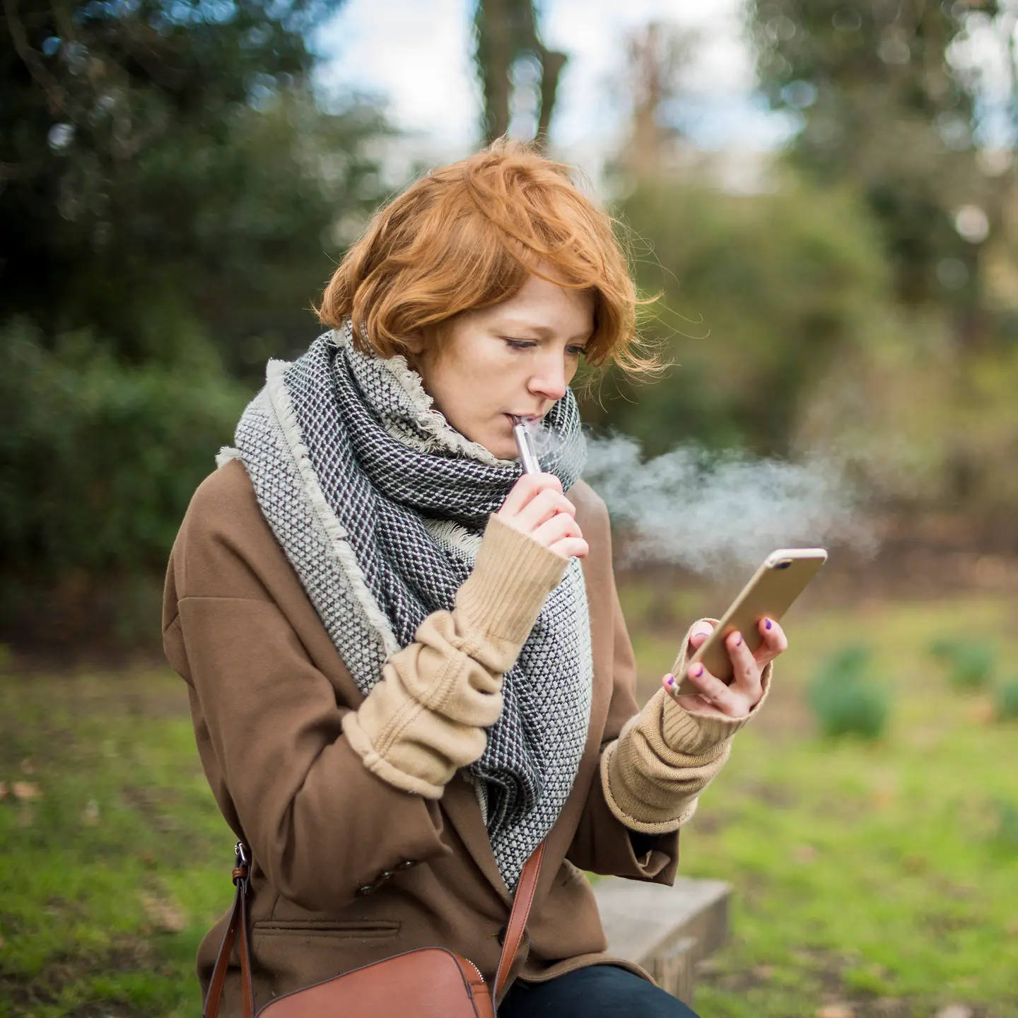 Eine junge Frau sitzt im Freien, schaut auf ihr Handy und raucht dabei eine E-Zigarette.