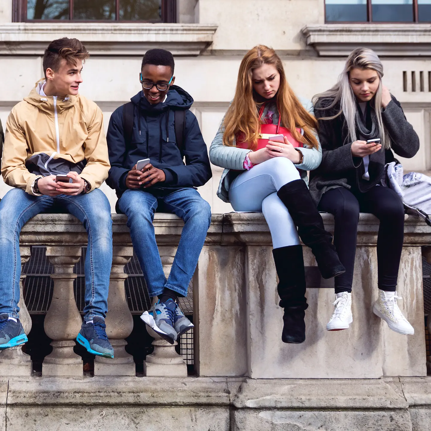 Eine Gruppe Jugendlicher sitzt auf einer Mauer und schaut auf ihr jeweiliges Smartphone.