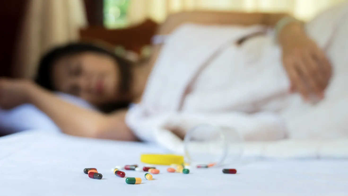 Eine Frau liegt schlafend im Bett, vor ihr liegt ein umgekippter Becher mit Tabletten.