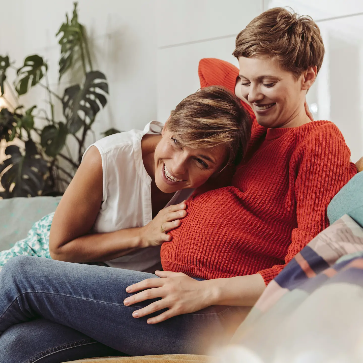 Eine schwangere Frau sitzt auf dem Sofa und ihre Partnerin legt ihre Hand und ihren Kopf auf den Babybauch