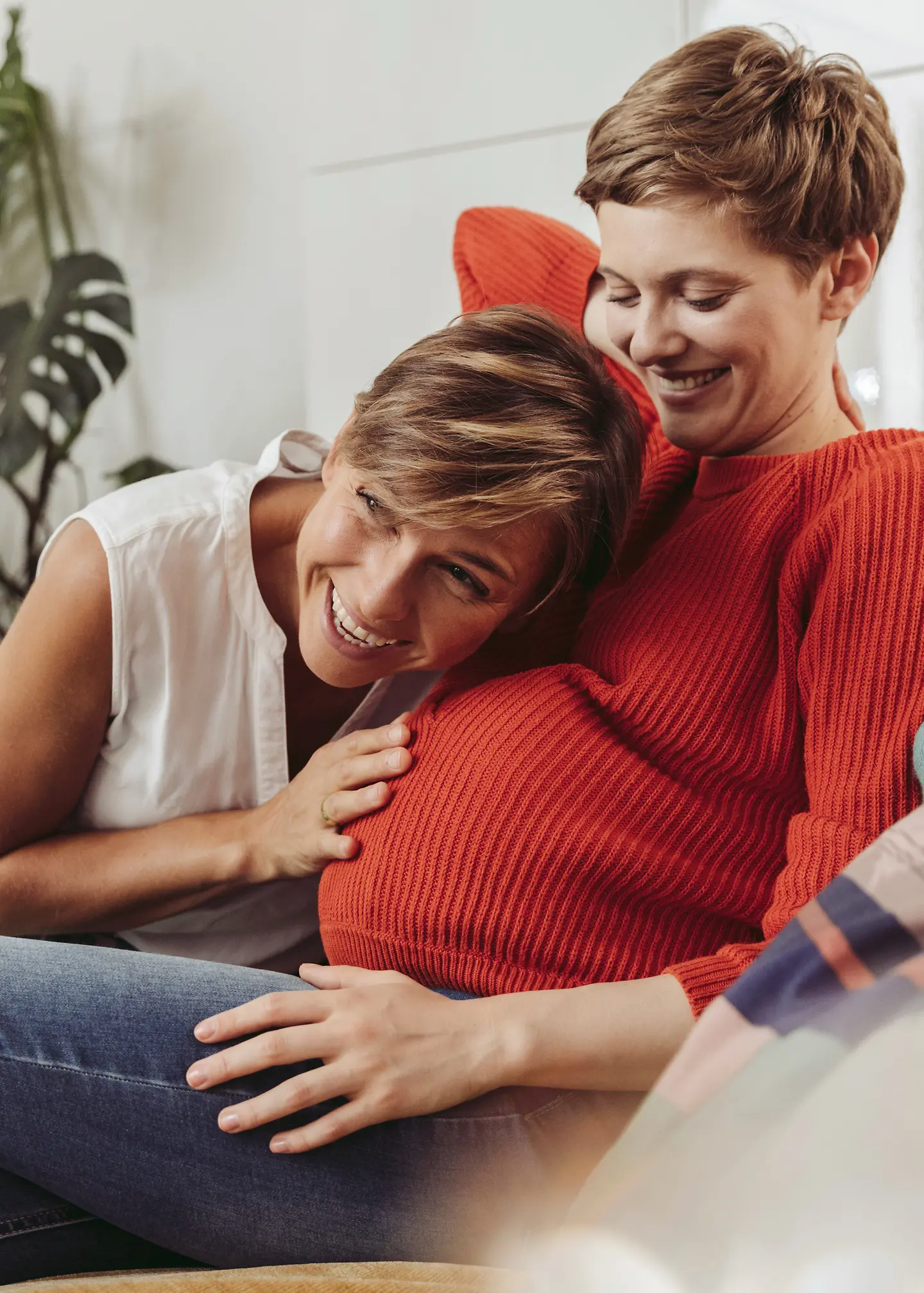 Eine schwangere Frau sitzt auf dem Sofa und ihre Partnerin legt ihre Hand und ihren Kopf auf den Babybauch