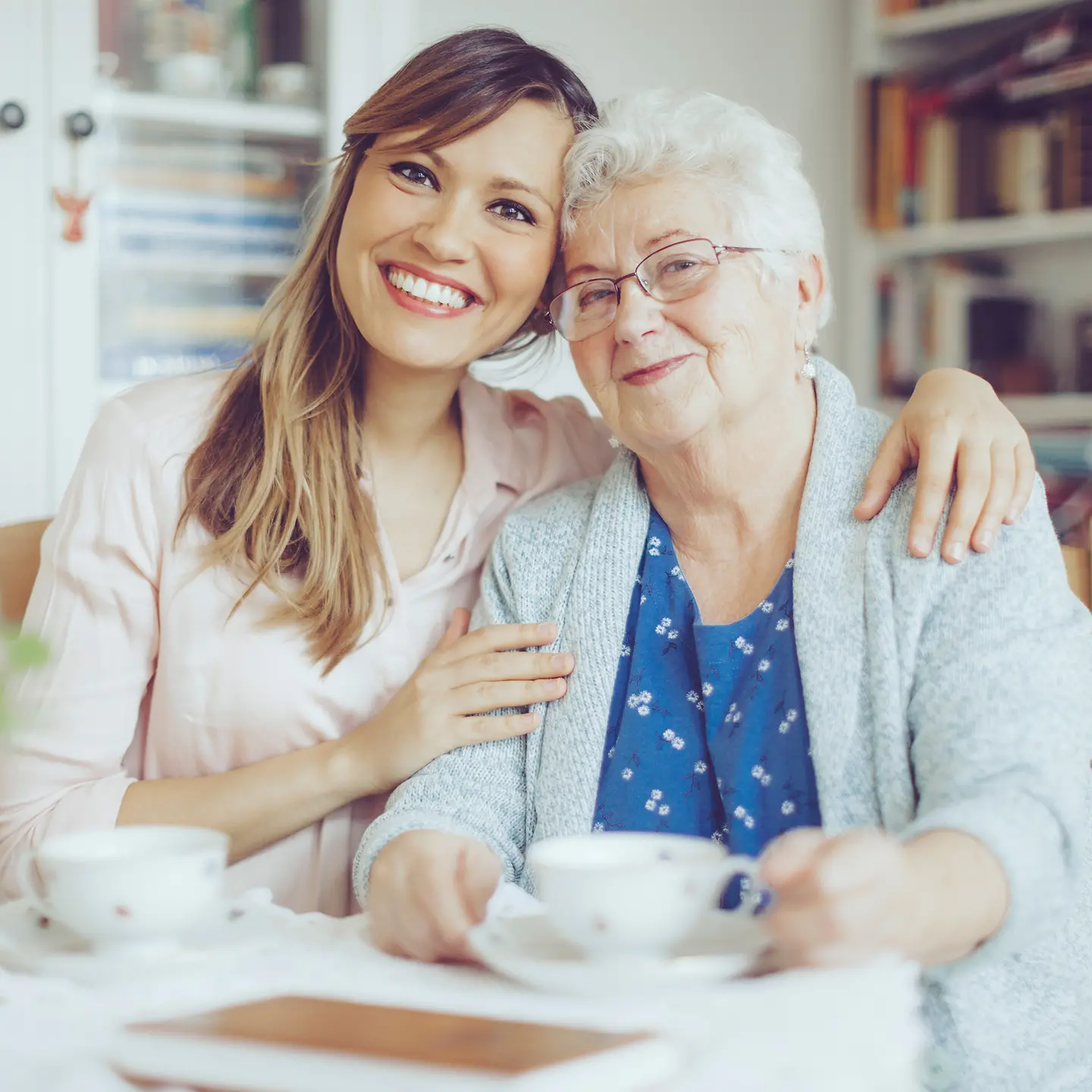 Alte Frau sitzt am Tisch und wird von junger Frau liebevoll umarmt