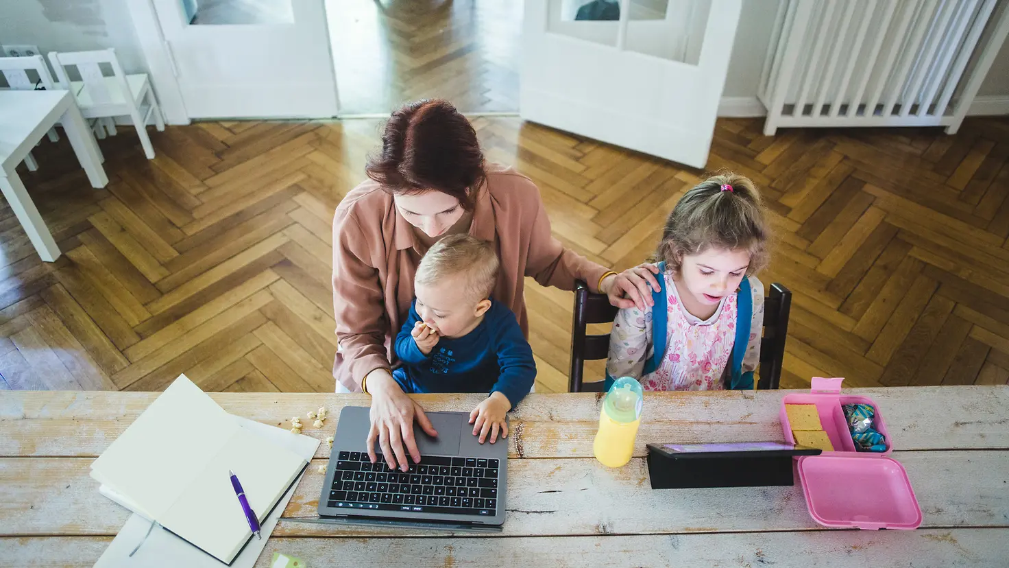 Das Bild zeigt eine am Laptop arbeitende Frau mit Baby auf dem Schoß und Kleinkind neben ihr.