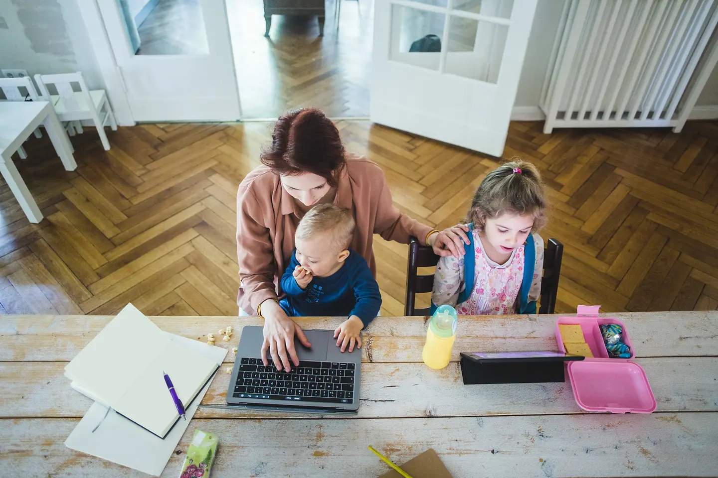 Das Bild zeigt eine am Laptop arbeitende Frau mit Baby auf dem Schoß und Kleinkind neben ihr.