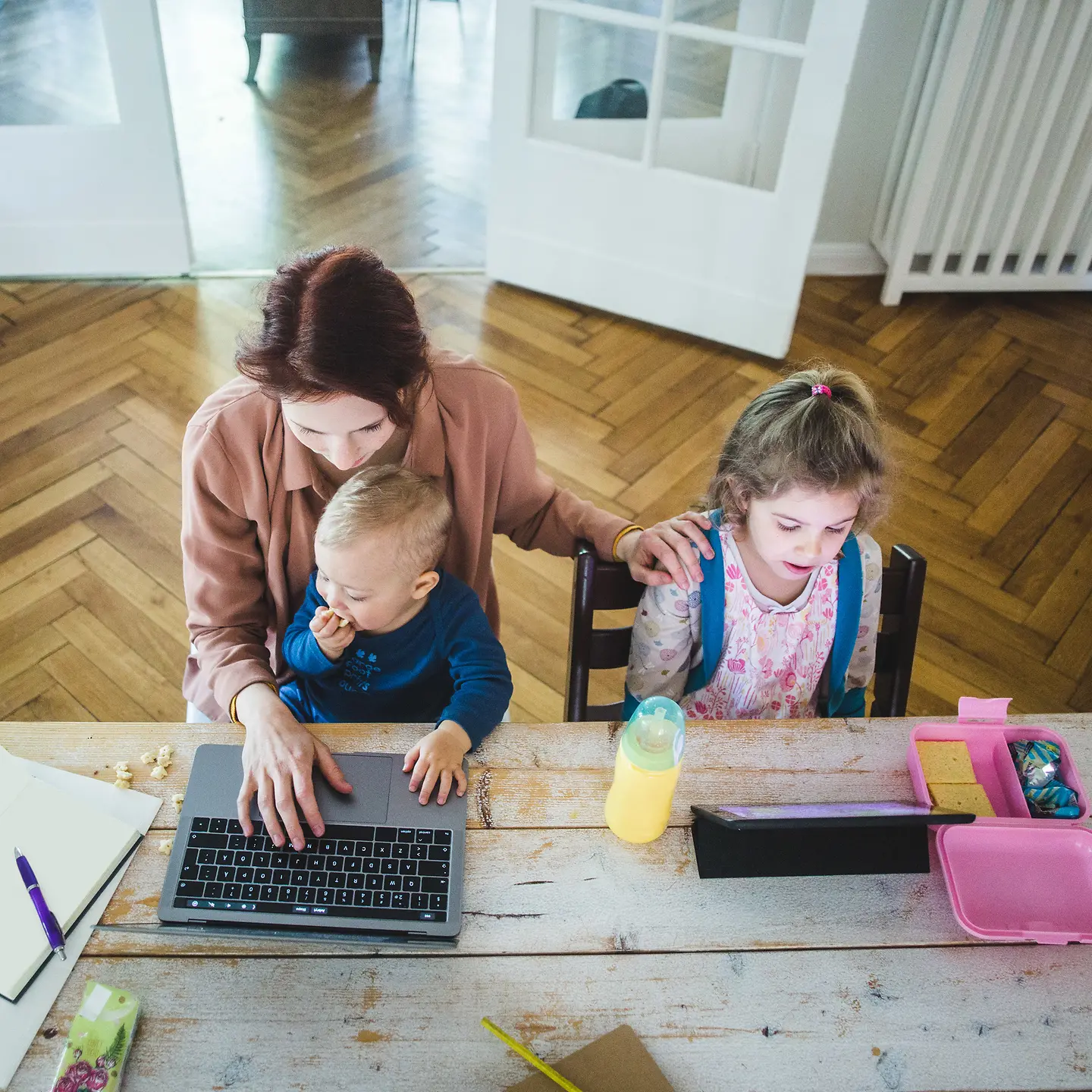 Das Bild zeigt eine am Laptop arbeitende Frau mit Baby auf dem Schoß und Kleinkind neben ihr.