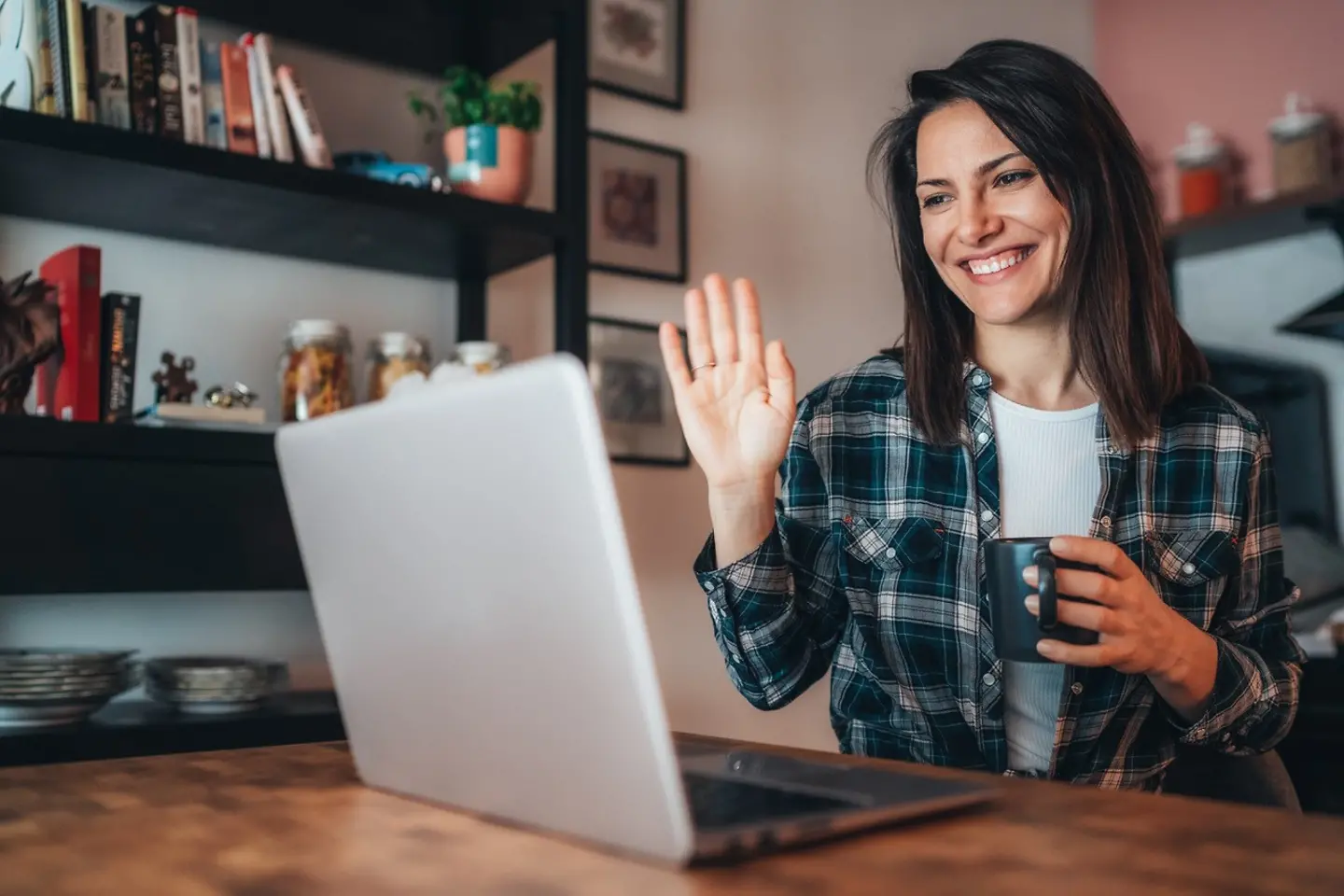 Eine lächelnde Frau sitzt an einem Schreibtisch vor einem Bildschirm und winkt in dessen Richtung.