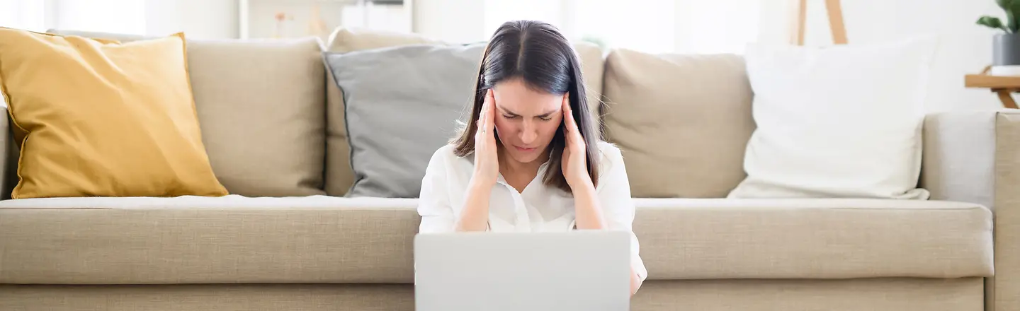 Frau sitzt mit Arbeitsutensilien und Kopfschmerzen auf dem Wohnzimmerfussboden