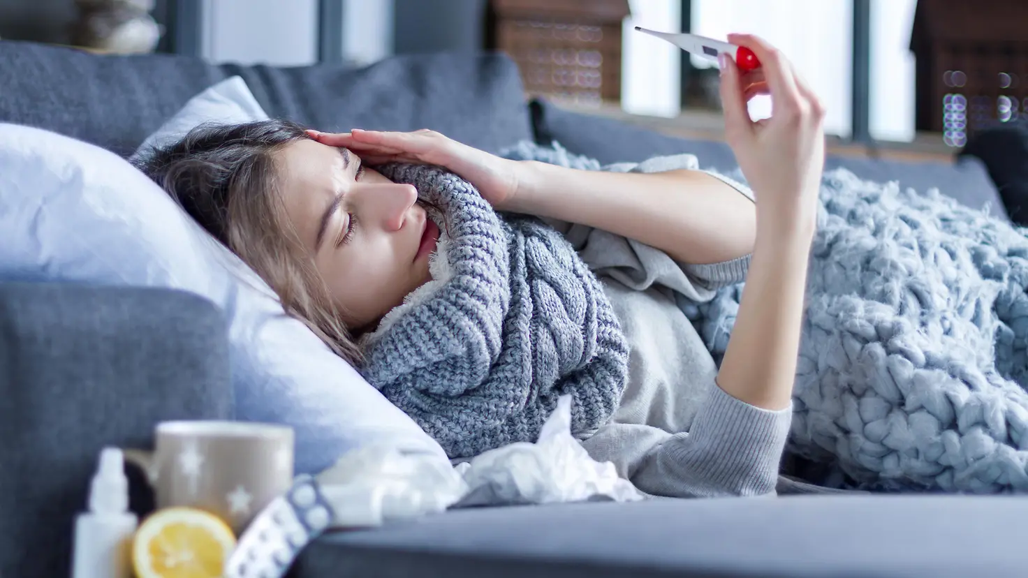 Eine Frau liegt mit Grippe auf dem Sofa, hält sich eine Hand an den Kopf und in der anderen ein Fieberthermometer.