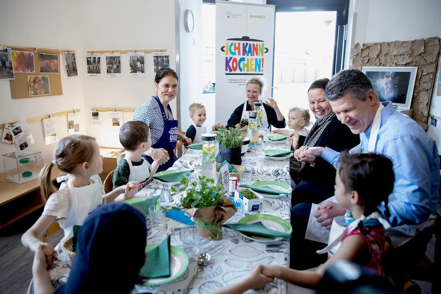 Kinder und Erwachsene sitzen am gedeckten Tisch und fassen sich an den Händen.