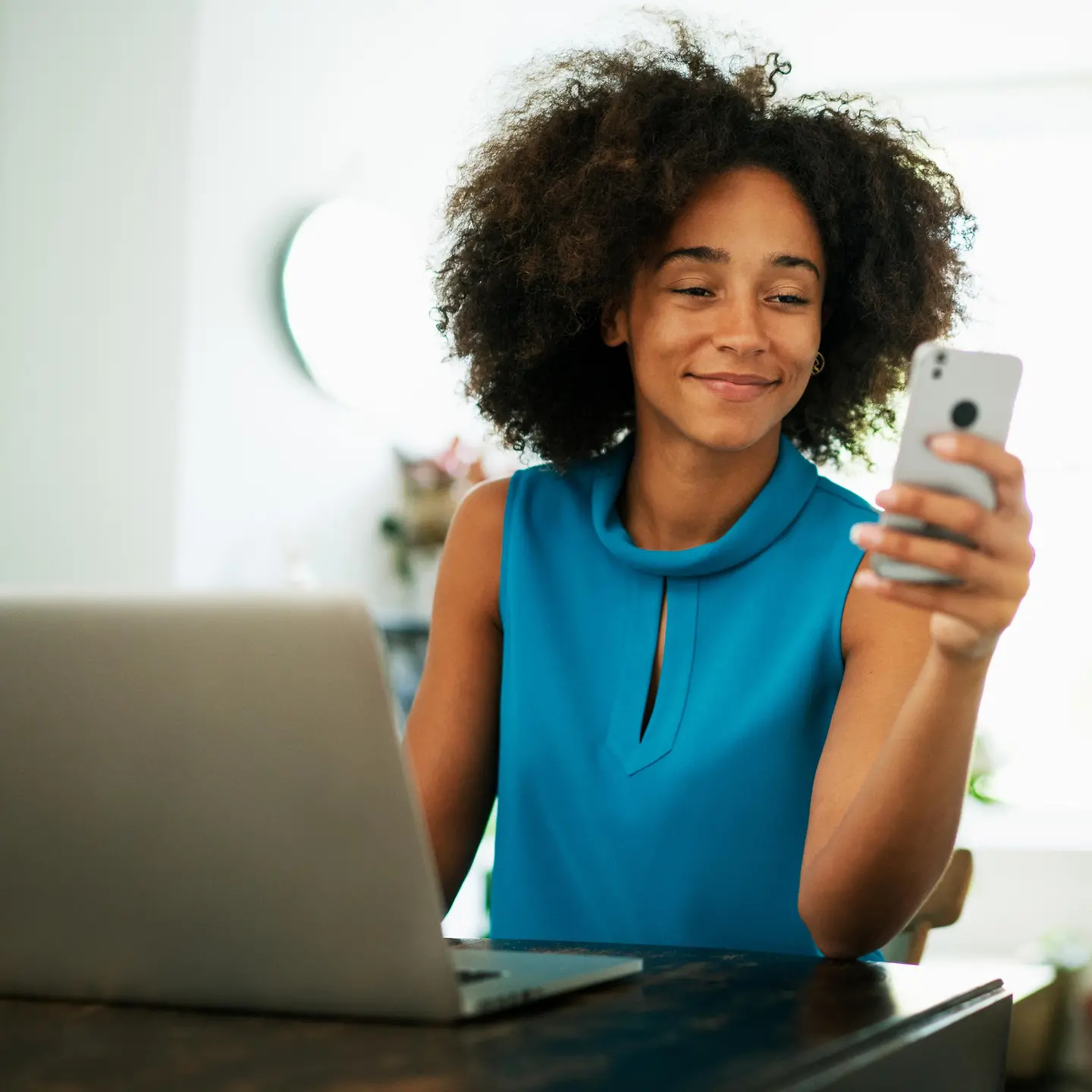  一位年輕女子坐在筆記本電腦旁，手裡拿著一部智能手機。