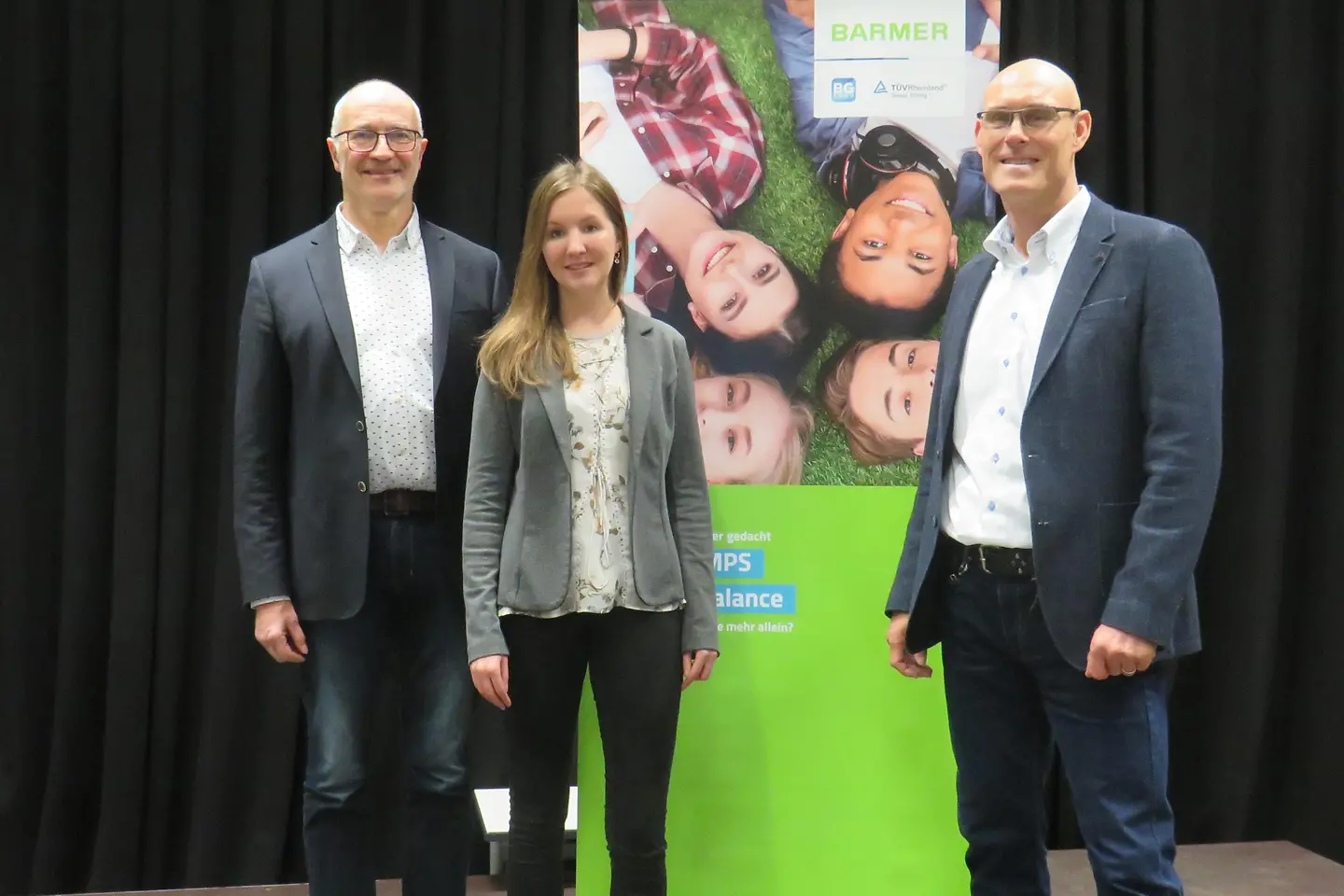 Schulleiter, Mitarbeiterin BG3000 und BARMER-Vertreter stehen vor einer Wand