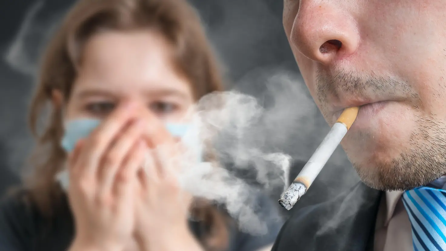 Dieses Bild zeigt einen Mann mit Zigarette im Mund, im Hintergrund hält eine Frau die Hände vor Mund und Nase