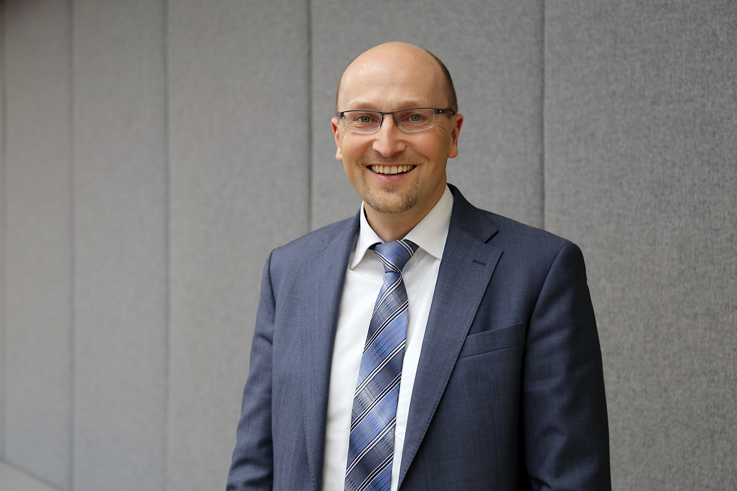 Ein Mann mit Glatze, Brille und Anzug lächelt in die Kamera