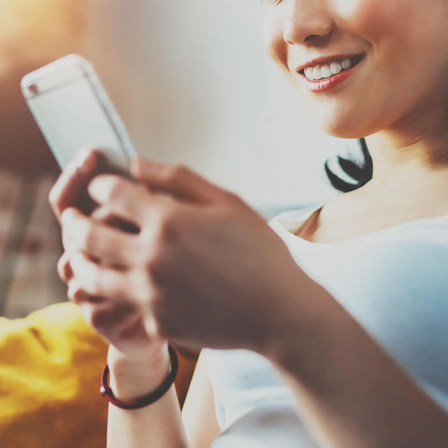 Eine junge Frau sitzt auf einem Sofa und schaut lächelnd auf ihr Smartphone in der Hand.