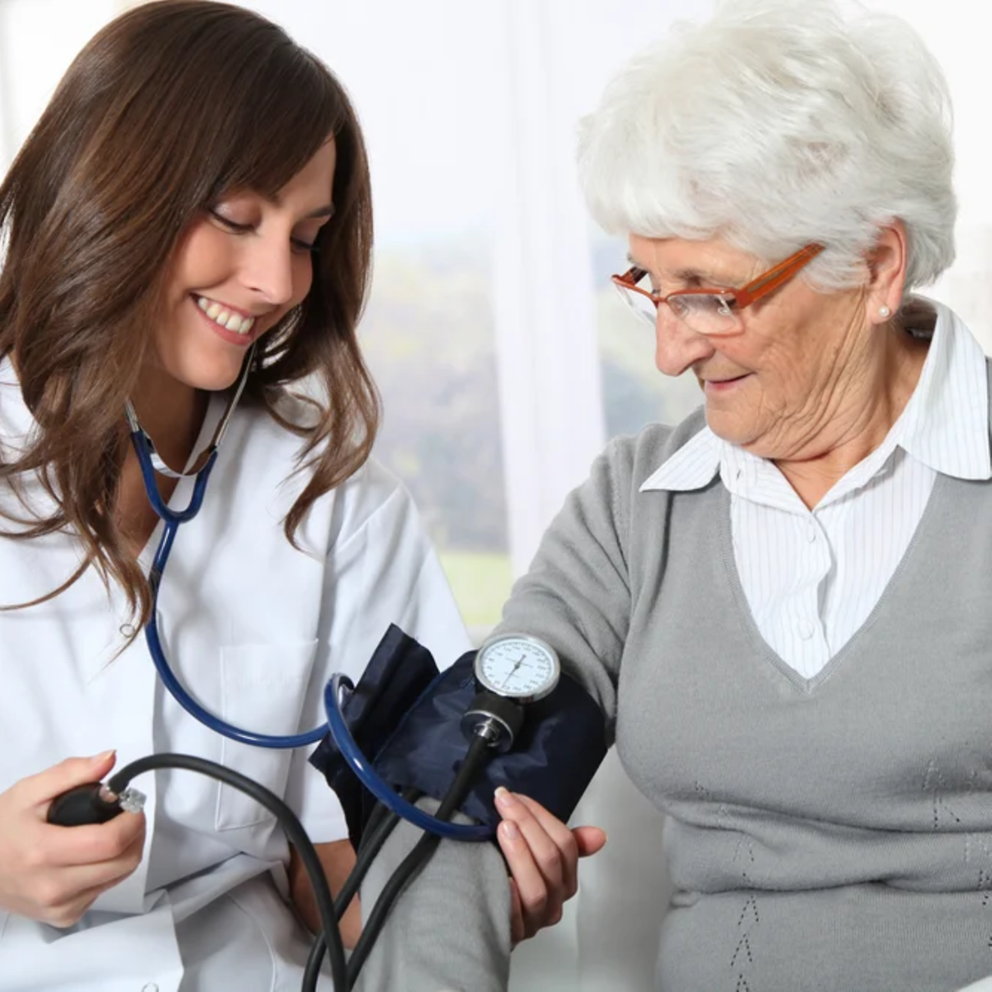 Eine Arzt-Schwester im weißen Kittel misst einer älteren Patientin mit weißen Haaren den Blutdruck.