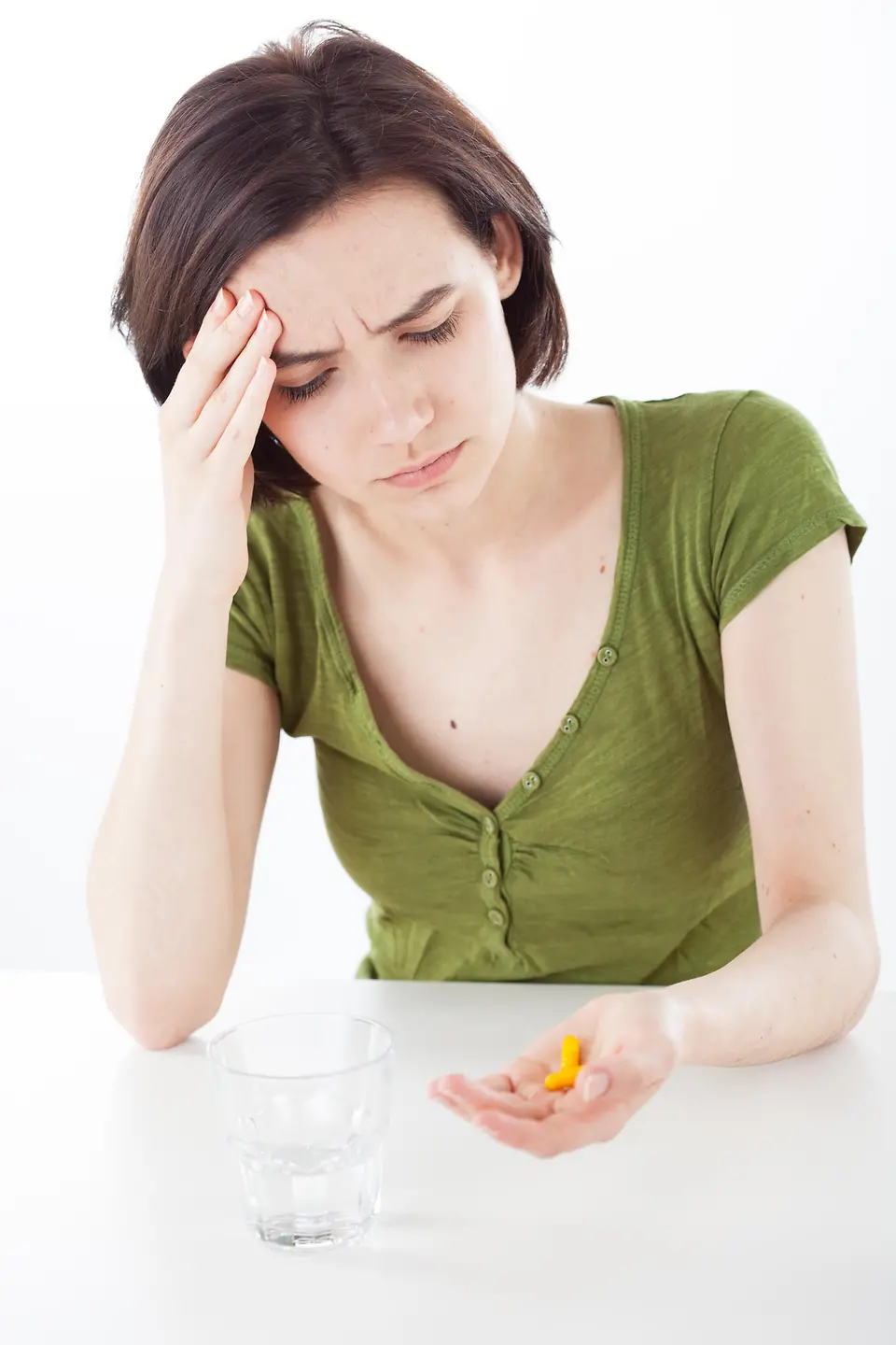 Eine Frau sitzt an einem weißen Tisch und hält in einer Hand Tabletten, die andere Hand fasst an die Stirn. 