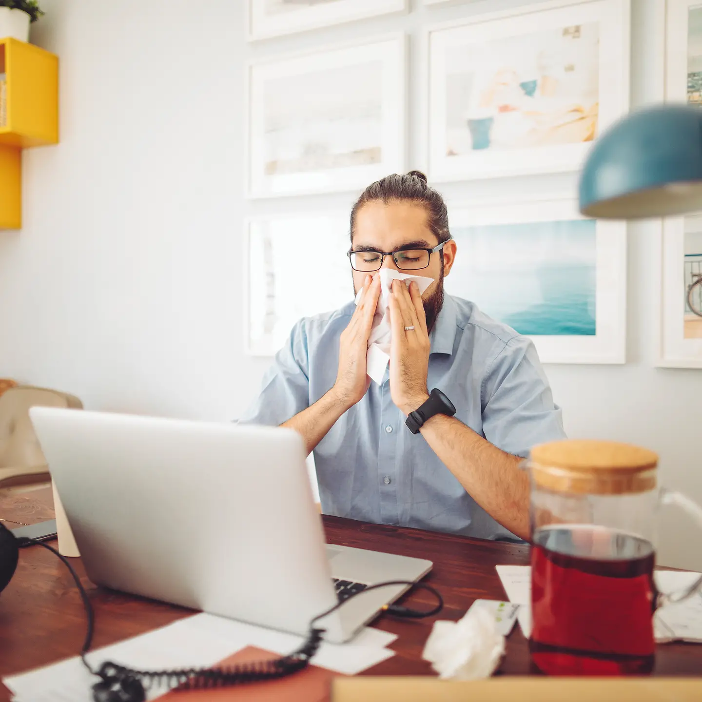 Ein junger Mann sitzt an einem Notebook im Homeoffice und putzt sich die Nase.