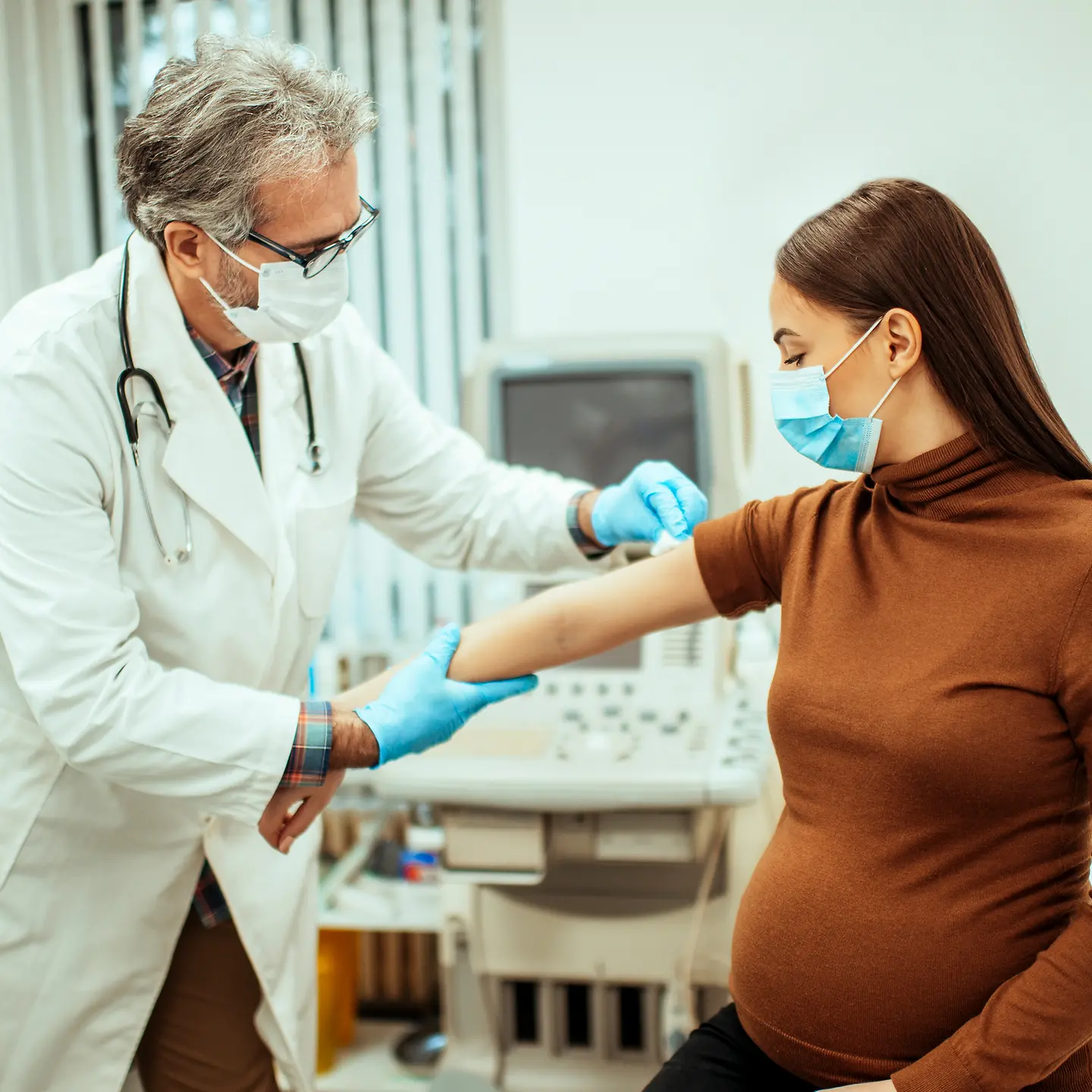 In einer Arztpraxis hält ein Arzt den Arm einer schwangeren Frau und desinfiziert eine Stelle am Oberarm für eine Impfung.